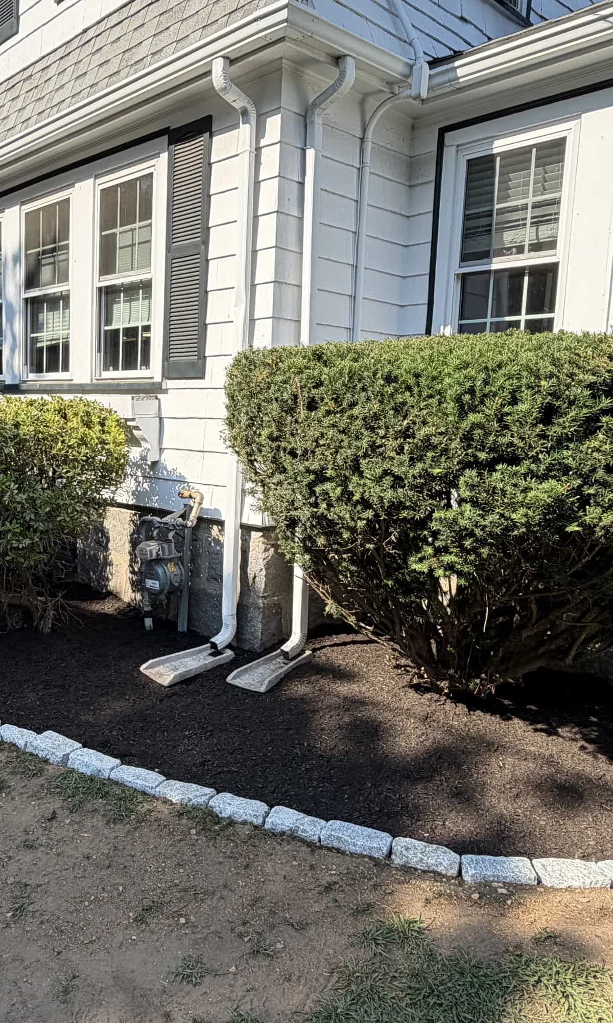Fresh dark mulch and trimmed shrubs along a granite stone border in front of a white house.
