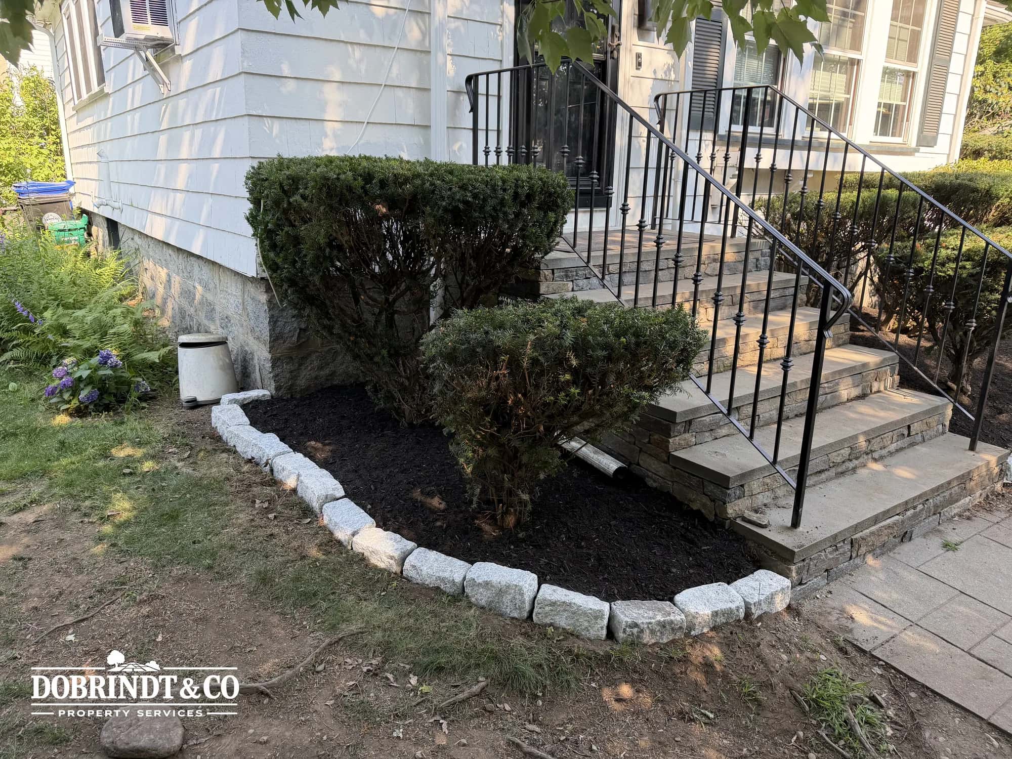Trimmed shrubs and a fresh mulch bed with granite edging beside front steps.