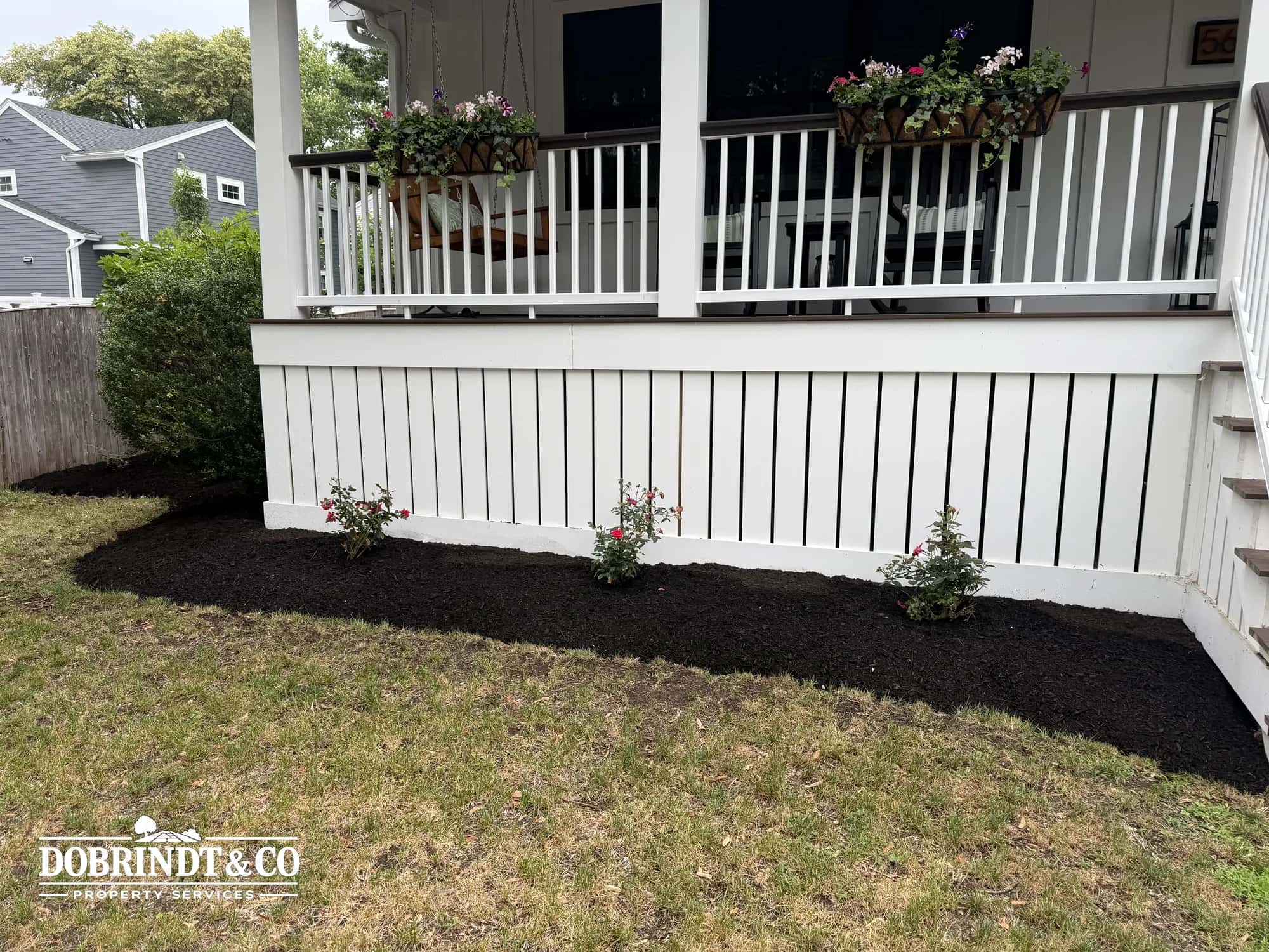 Black mulch and flowering plants beneath a porch with a white railing.