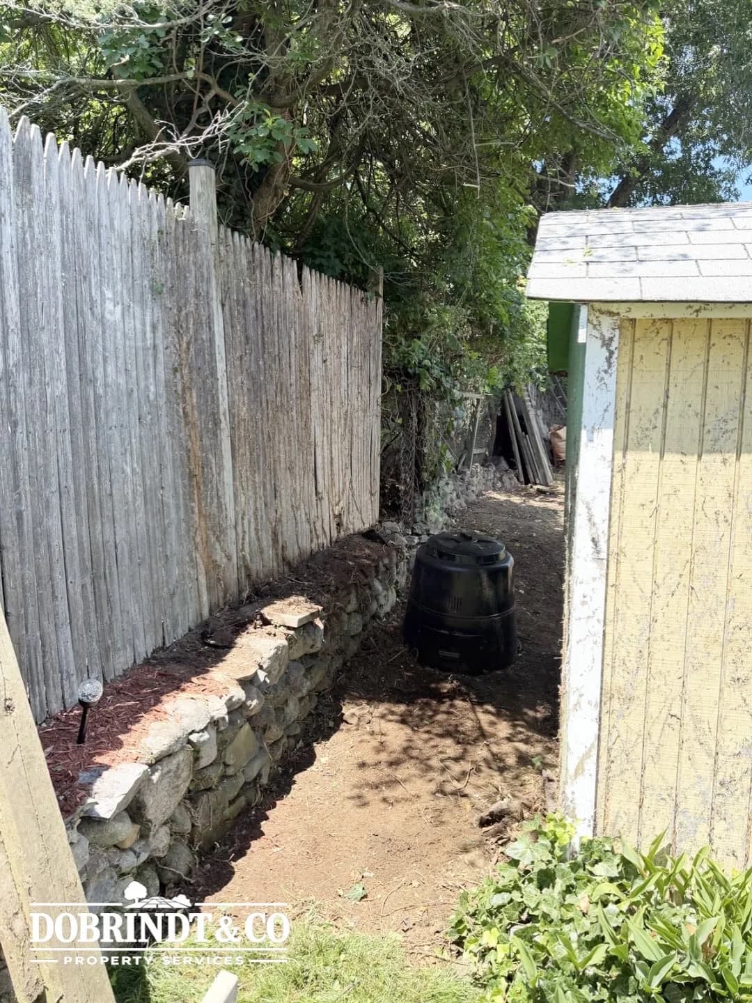 Backyard enclosed by a wooden fence with a compost bin along a stone-lined path and a shed in the background.