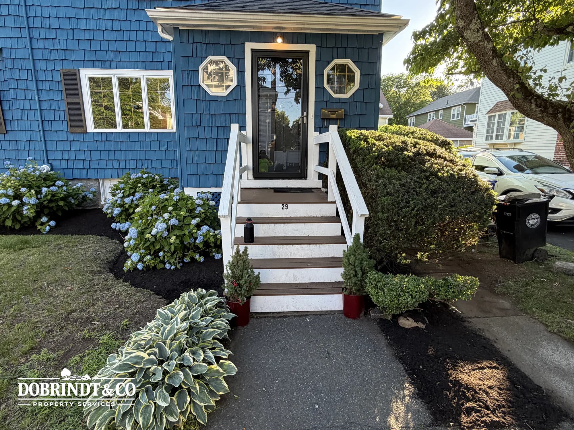 Exterior of a blue-shingled house with a tidy front yard of hydrangeas and white entry stairs.