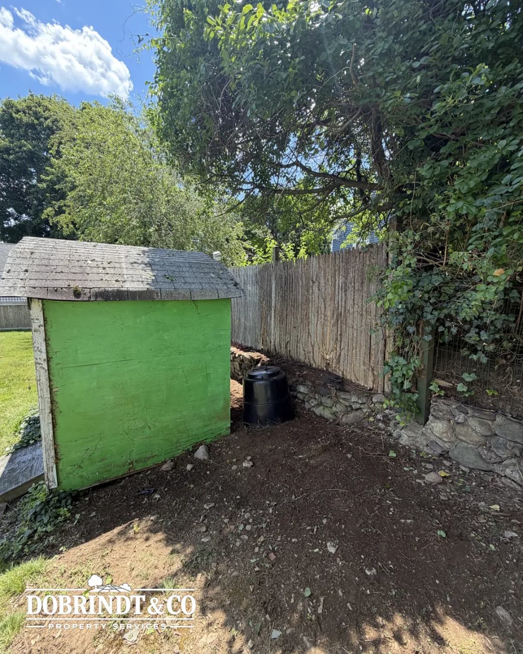 Small green shed in a grassy yard with a wooden fence and trees in the background.