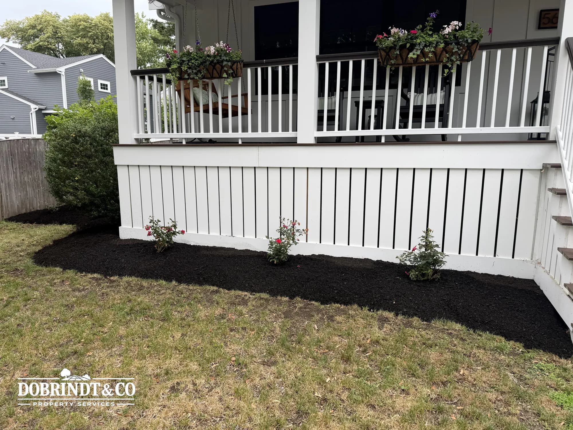 Well-maintained porch with flowering plants in black mulch and hanging planters on a white railing.